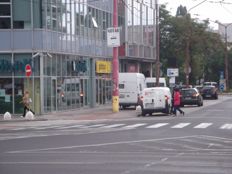 Biskupské klobúky na zabránenie parkovania na chodníku a žltá čiara. Napriek tomu sa parkuje aj na žltej čiare (treba však priznať, že zásobovanie tu nemá kde legálne zastať). A parkuje sa aj na chodníku, kvôli biskupským klobúkom však autá jazdia cez priechody pre chodcov. V odraze vidno dodávku, ktorá cúva cez priechod pre chodcov pri vjazde do garáže. Na legálnosť sa ani nemá zmysel pýtať, čo však policajti na bezpečnosť takýchto manévrov?